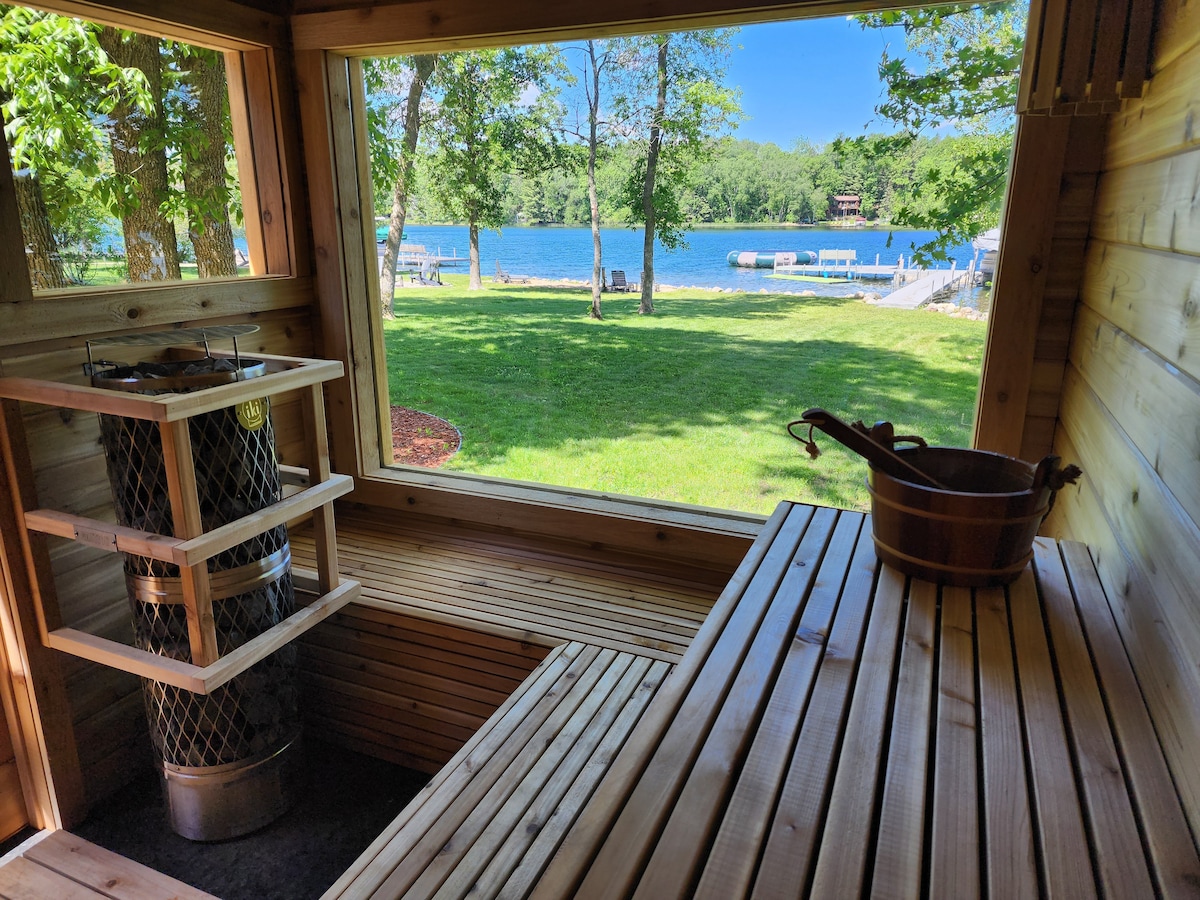 A wooden sauna is visible through a large window, offering a view of the lake and surrounding greenery. The interior is lined with wooden benches, and a metal stove sits beside a wooden bucket, providing a tranquil atmosphere for relaxation.