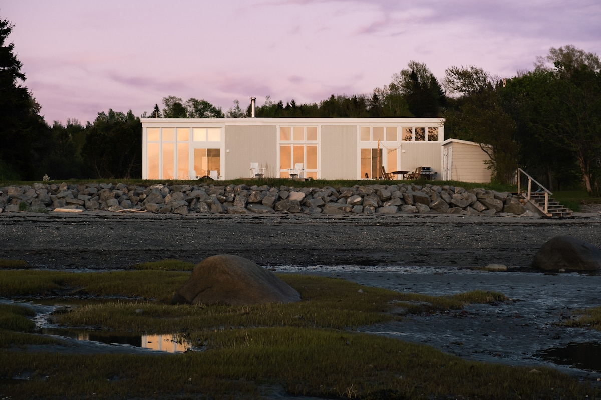 A modern seaside residence is captured at dusk, featuring large windows that reflect the warm hues of the sunset. The property is situated near the water, with a rocky shore and lush greenery visible in the foreground.
