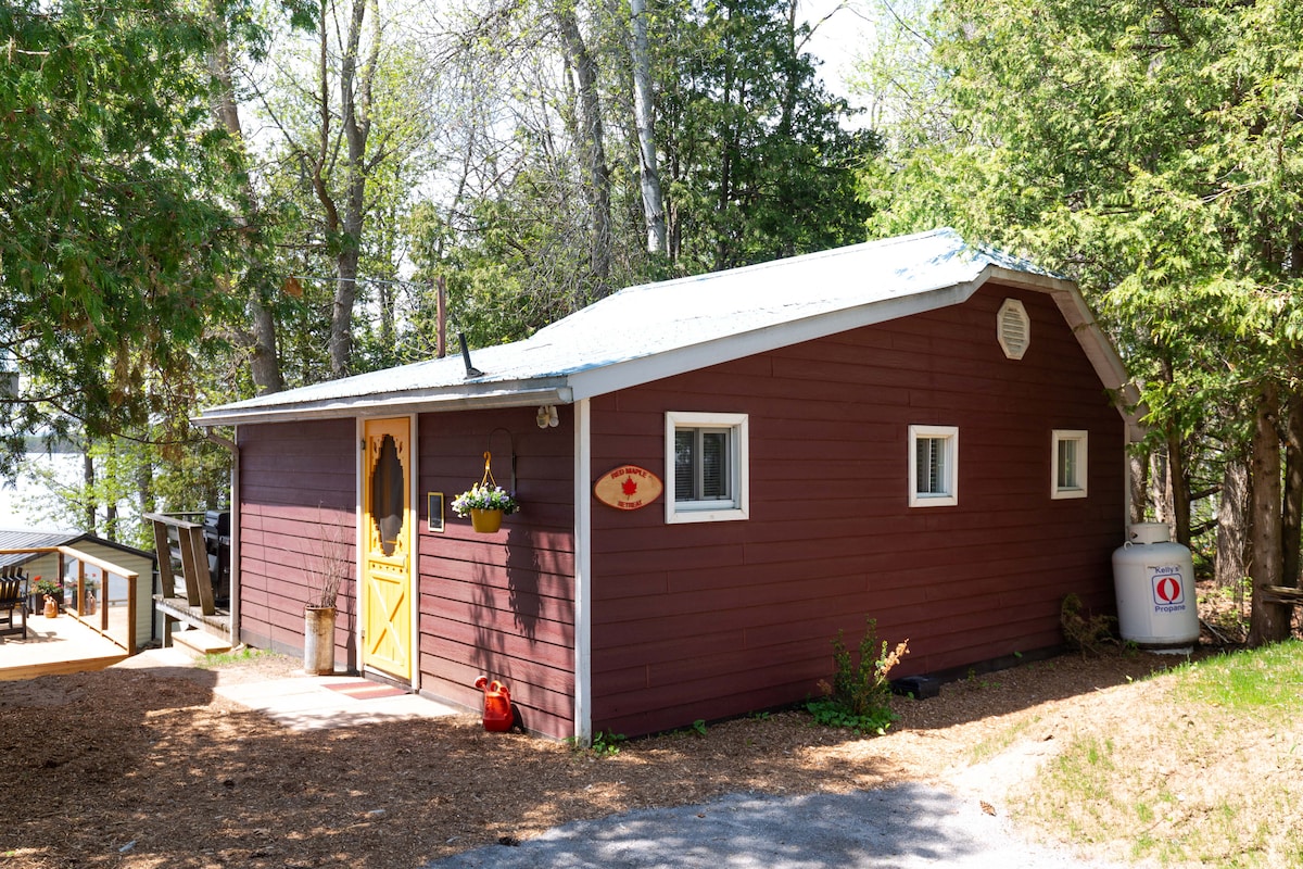 The exterior of a charming lakefront cottage features a warm red facade and a yellow door, inviting guests to enter. Surrounding greenery and a gravel path lead toward the entrance, while nearby outdoor seating is visible by the lake, providing a serene ambiance.