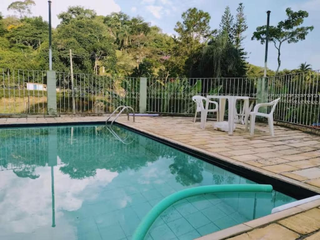 A tranquil outdoor space features a suspended swimming pool surrounded by a stone patio. Two white chairs and a table are positioned nearby, complemented by lush greenery visible in the background, creating a peaceful atmosphere.