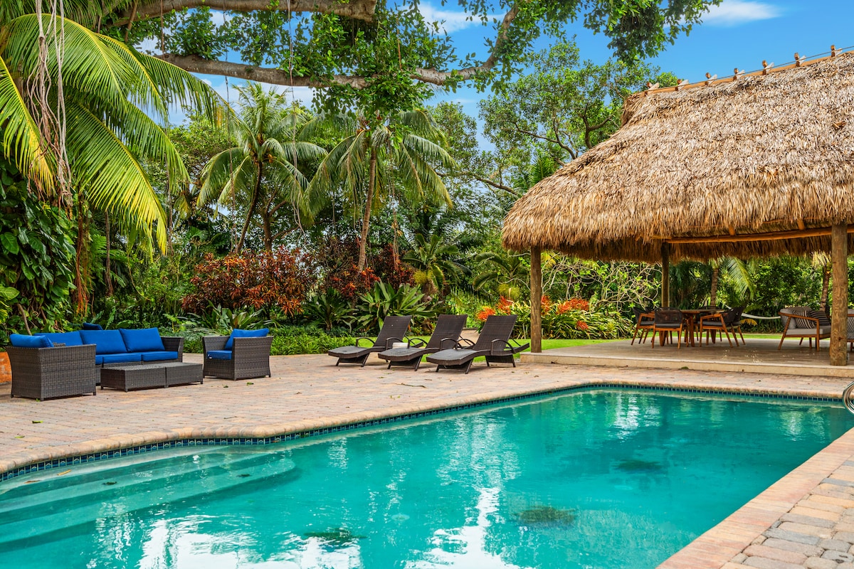A tranquil pool area is surrounded by lush tropical gardens, featuring a thatched-roof gazebo and ample seating. Comfortable lounge chairs line the poolside, inviting relaxation under the sun. The clear water reflects the greenery, creating a serene outdoor setting.