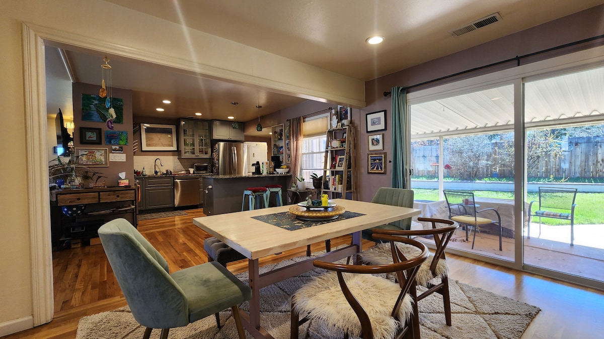 An open living space is visible, featuring a wooden dining table with a centerpiece. Surrounding the table are chairs in varying styles. The kitchen area is equipped with modern appliances and cabinets, while large sliding glass doors offer a view of an outdoor patio and landscaped yard.