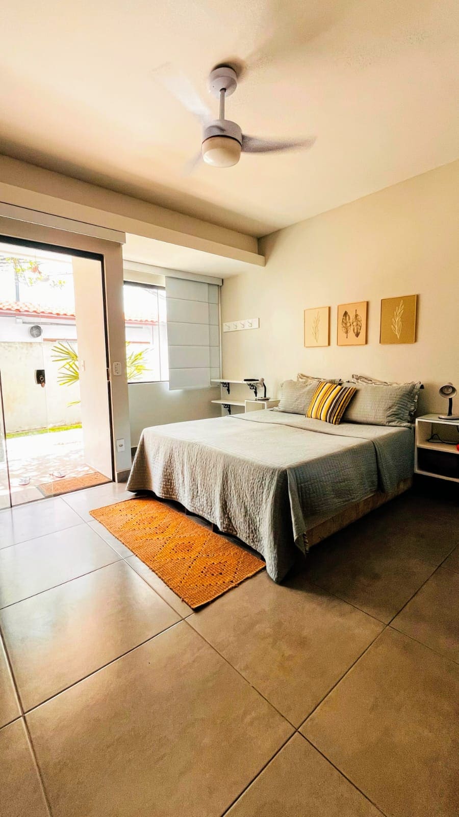 A bright bedroom features a spacious bed adorned with a neutral-toned bedspread and decorative pillows. Natural light enters through a glass door leading to an outdoor space, complemented by light-colored wall art. A small bedside table and rug add to the room's comfort.