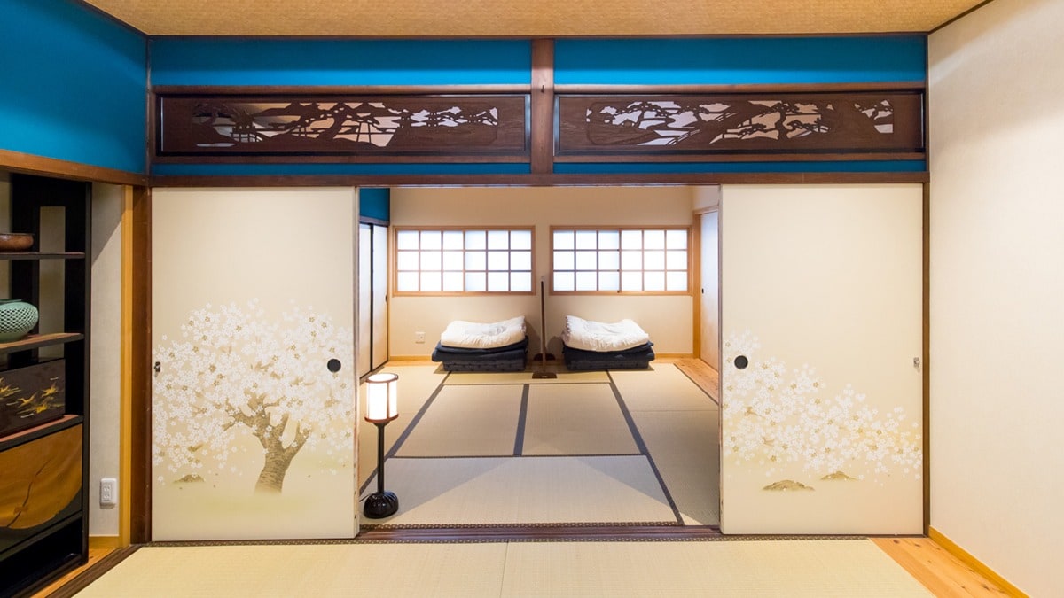 A traditional Japanese room features sliding doors adorned with cherry blossom artwork, leading to a second room illuminated by natural light from small windows. Light wooden flooring complements the tatami mats, while a soft ambient lamp provides additional warm light.