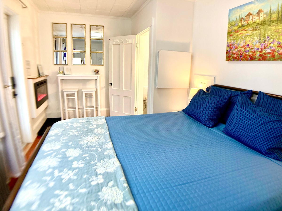 A cozy studio room features a spacious bed with a blue quilt, accent pillows, and hardwood floors. A fireplace is visible, along with a neatly arranged seating area. Light enters through a nearby window, illuminating a mounted artwork of a colorful landscape.
