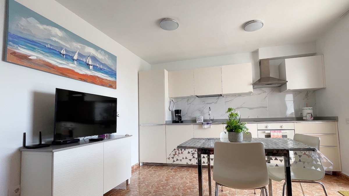 A modern kitchen area showcases sleek cabinetry with a marble backsplash. A dining table with light chairs is positioned nearby, complemented by a decorative plant. A television is displayed on a side cabinet, providing a functional and stylish space.