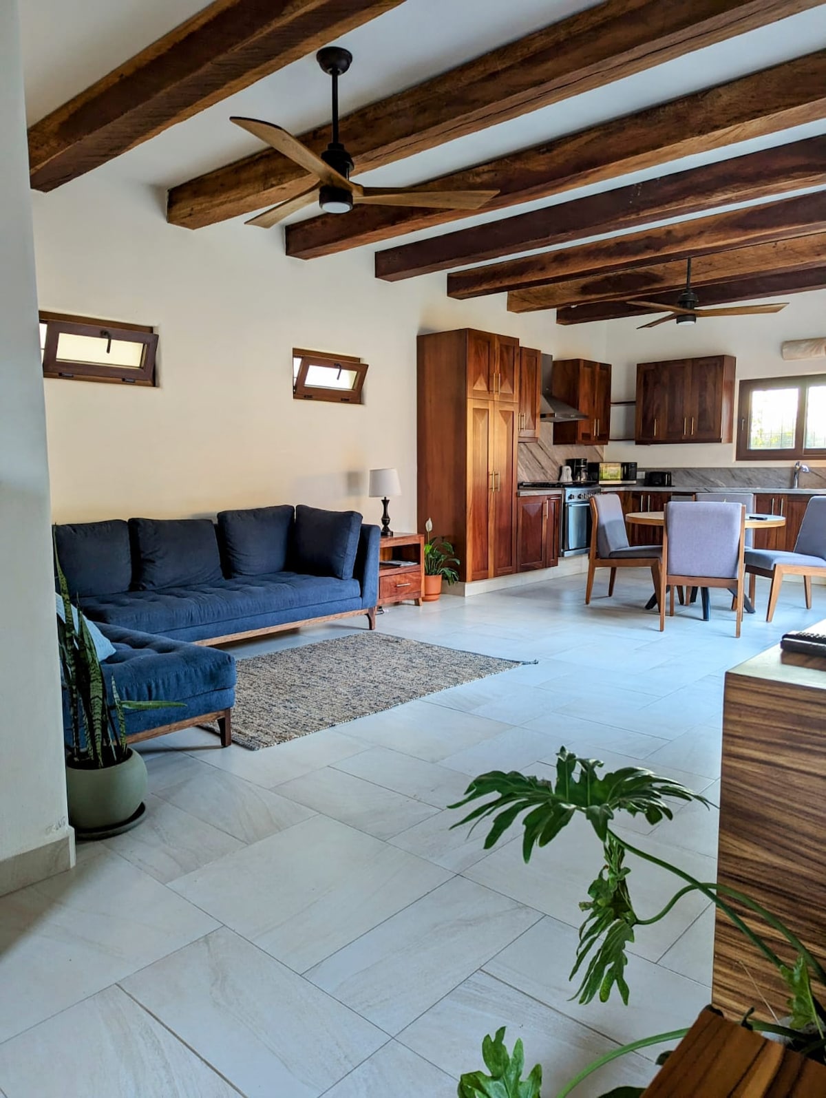 A light-filled living area is presented, featuring a navy blue sofa paired with a small side table and a lamp. Wooden beams accentuate the ceiling, while a dining table with four chairs is positioned near the kitchen. Indoor plants add a touch of greenery.