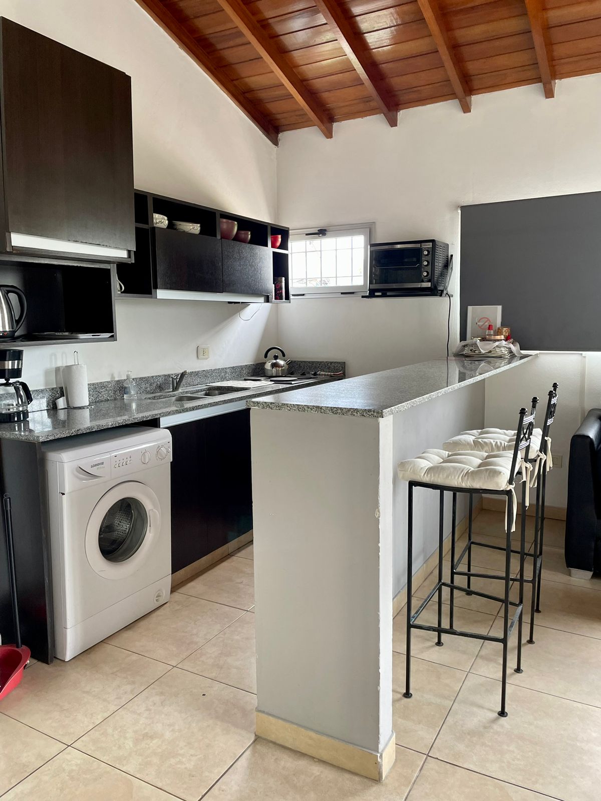 The kitchen area features modern cabinetry and countertop space, equipped with an electric oven, microwave, coffee maker, and blender. A washing machine is located beneath the countertop. Bar stools provide seating at the breakfast bar, enhancing functionality in the space.