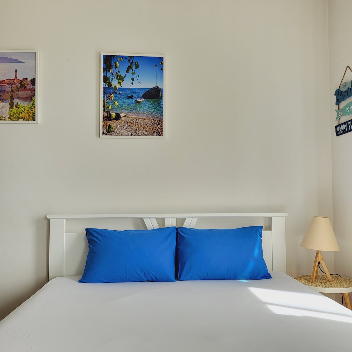 A bright sleeping area is highlighted by a bed with two blue pillows against a white headboard. Framed coastal images adorn the wall, while a small lamp and a natural fiber accent add warmth to the space. Light filters in, creating a cheerful atmosphere.