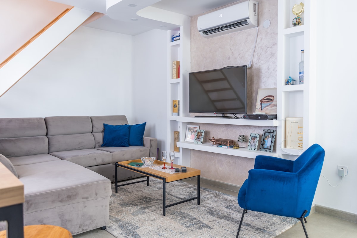 A modern living area features a spacious gray sectional sofa with blue accent pillows. A wooden coffee table is positioned in front, alongside a patterned rug. A wall-mounted television is integrated with tasteful shelving, which displays decorative items and books, contributing to the contemporary design.