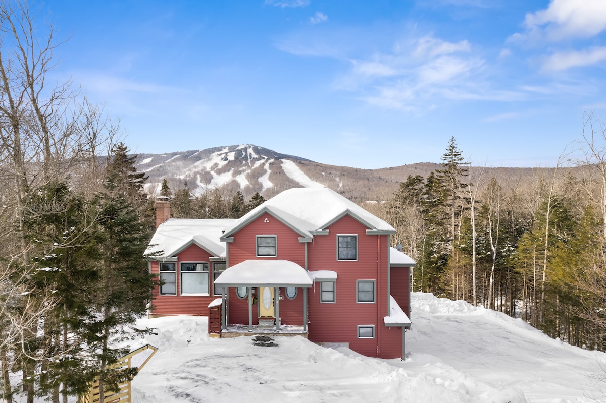 The exterior of the house is shown in a snowy landscape, complemented by a backdrop of Mount Snow. The structure features a red façade with large windows and a welcoming front porch, framed by trees that highlight the serene winter setting.