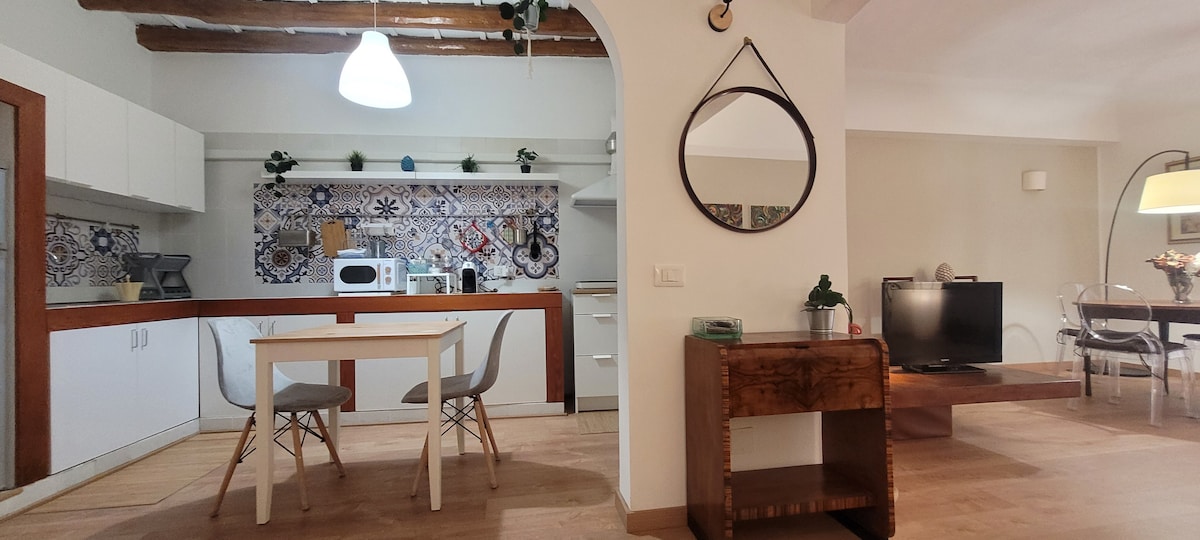 A kitchen area is visible, featuring white cabinetry and decorative tiled backsplash. A dining table with four chairs is present, complemented by a modern light fixture overhead. A cozy living area is seen in the background with a television, enhancing the spacious layout.