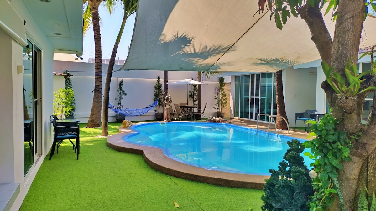 A swimming pool is surrounded by a green lawn and palm trees, creating a serene outdoor area. Comfortable seating is arranged beside the pool, while shaded spaces are provided by a large canopy. The pool area is complemented by tropical plants, enhancing the tranquil setting.