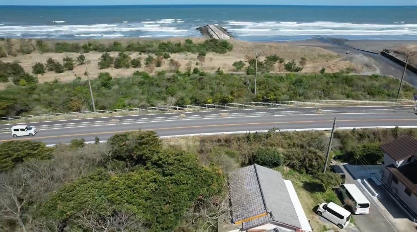 An aerial view captures the coastline, showcasing waves breaking along the shore. A road runs parallel to the beach, bordered by greenery. The image includes several vehicles parked along the roadside, providing a sense of accessibility to the scenic area.
