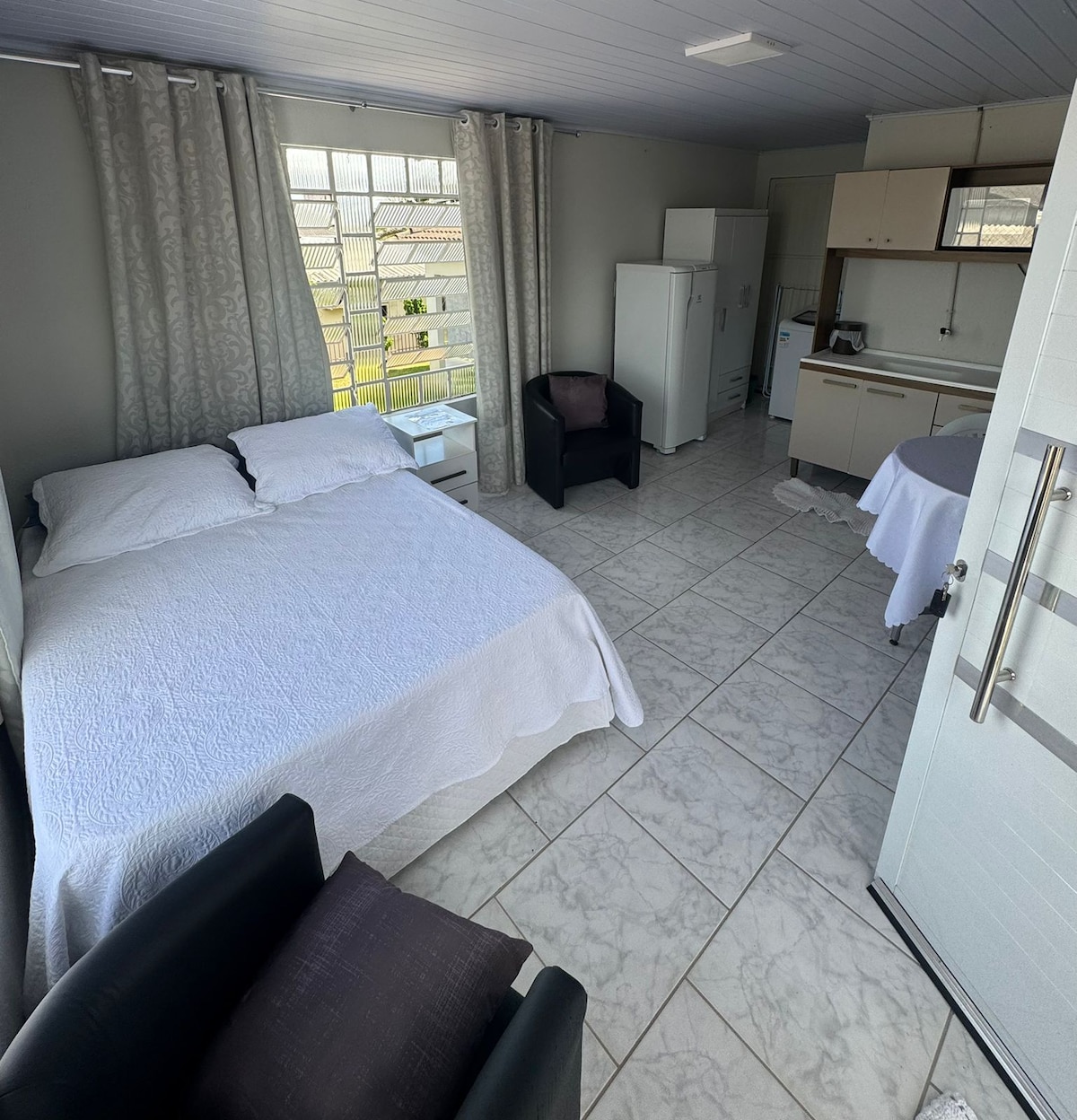 A well-lit room contains a spacious bed covered with a plain white quilt, flanked by two black chairs. A compact kitchenette is visible in the background, accompanied by a refrigerator and light-colored cabinetry. Large windows offer views and allow natural light to fill the space.