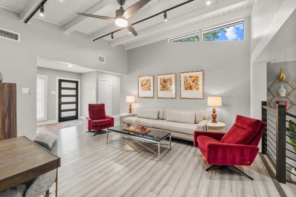 The living area features a light-colored sofa complemented by red accent chairs. Natural light filters through large windows, illuminating the space. A glass coffee table sits in the center, surrounded by tasteful artwork that adorns the walls, and a wooden dining table is positioned nearby.