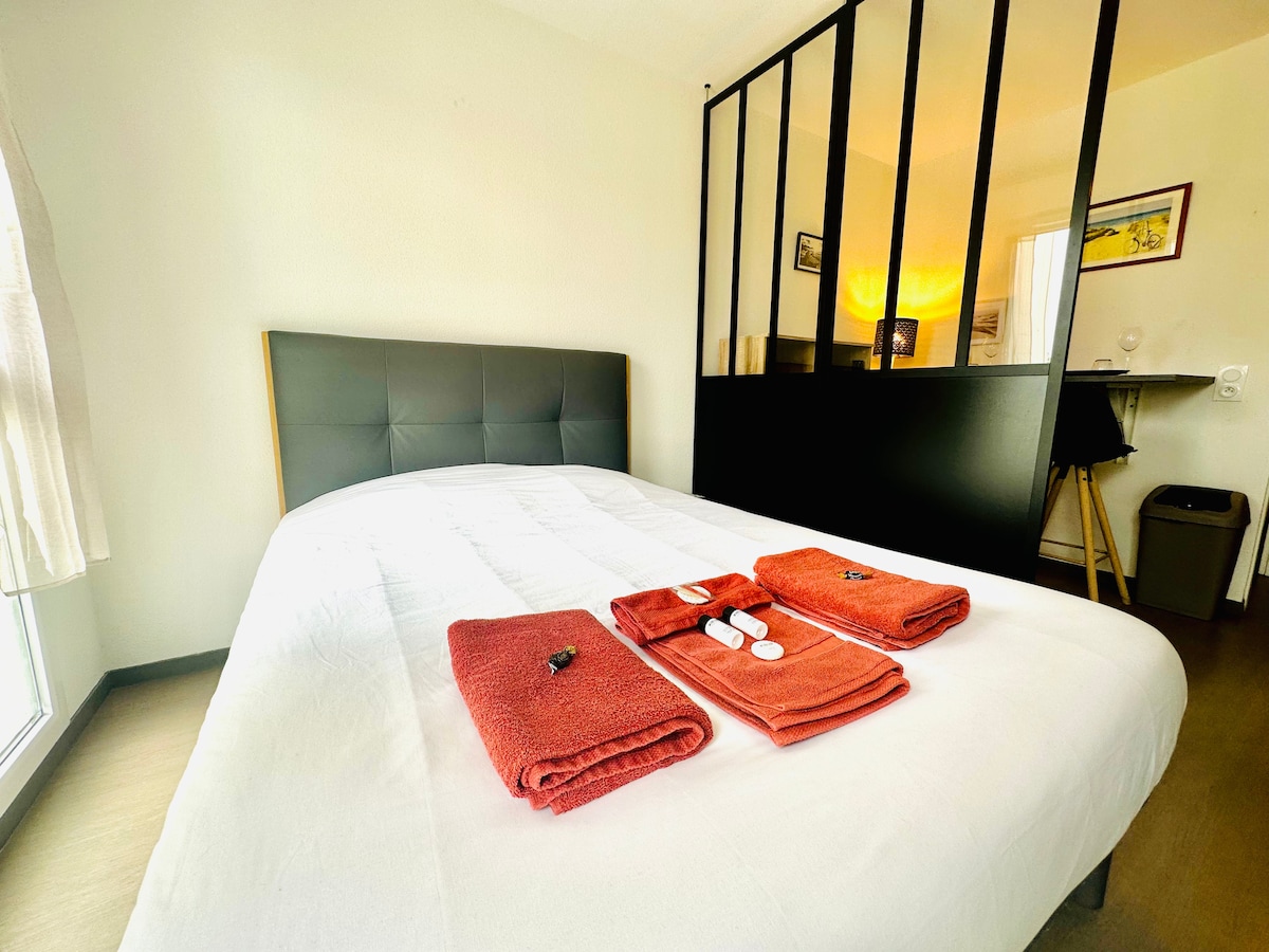 The sleeping area features a spacious bed with a neatly arranged white duvet. Two sets of bright orange towels are displayed at the foot of the bed alongside toiletries. A sleek partition divides this area from the kitchenette, creating an open yet cozy atmosphere.