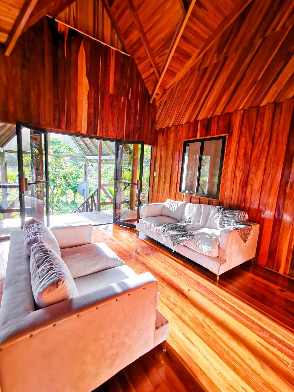 A bright living area features large glass doors that open to the outdoor space, allowing natural light to fill the room. The warm wooden walls and polished wooden floors enhance the inviting atmosphere. Two comfortable sofas are arranged for relaxation, complemented by a chic throw.