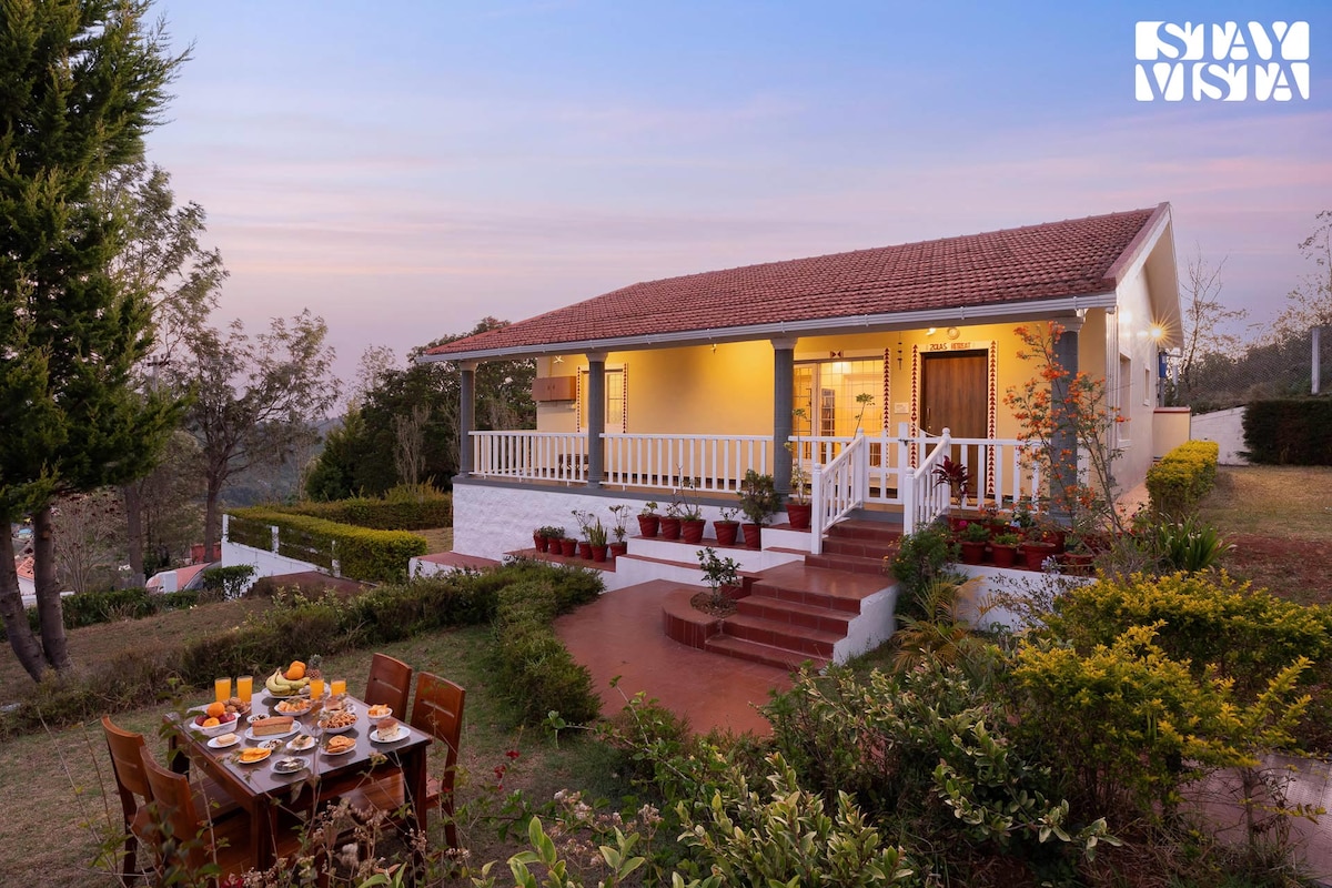 A charming villa is set against a beautiful twilight sky, surrounded by lush greenery. A dining table is arranged outside with breakfast items, while the villa features a spacious porch with white railings and potted plants. Steps lead up to the entrance, enhancing accessibility.