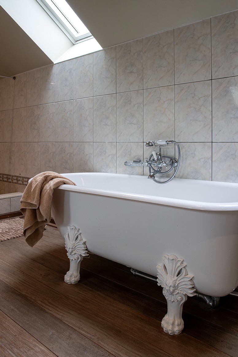 A freestanding bathtub is positioned in a softly lit bathroom, featuring decorative claw feet. A beige towel rests casually on the tub's edge, while tiled walls in muted tones provide a serene backdrop. Natural light filters in through a skylight above.