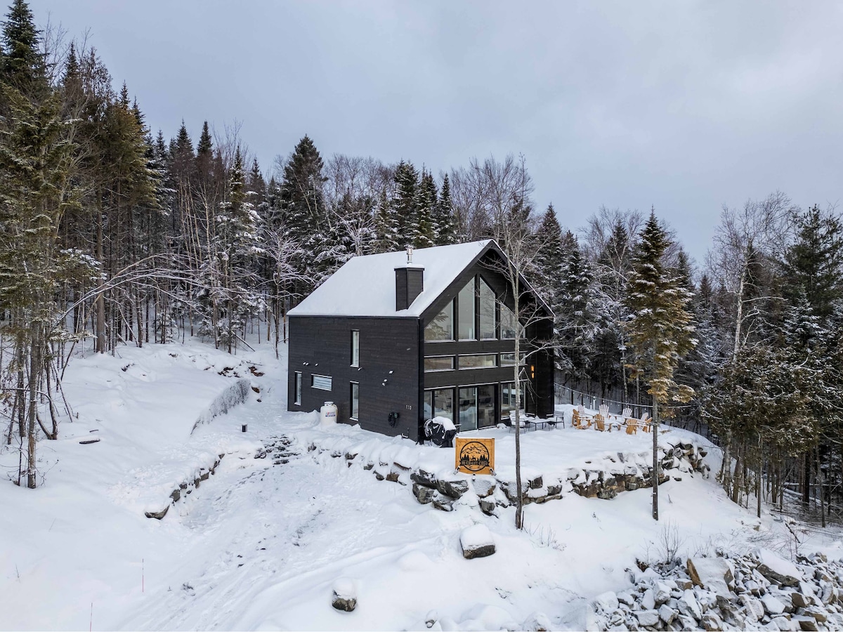 The chalet is situated on a snowy hillside, surrounded by trees. Large windows offer views of the winter landscape. Outdoor seating is visible on the terrace, contributing to the natural aesthetic. The entrance is marked by a wooden sign, indicating the property's welcoming presence.