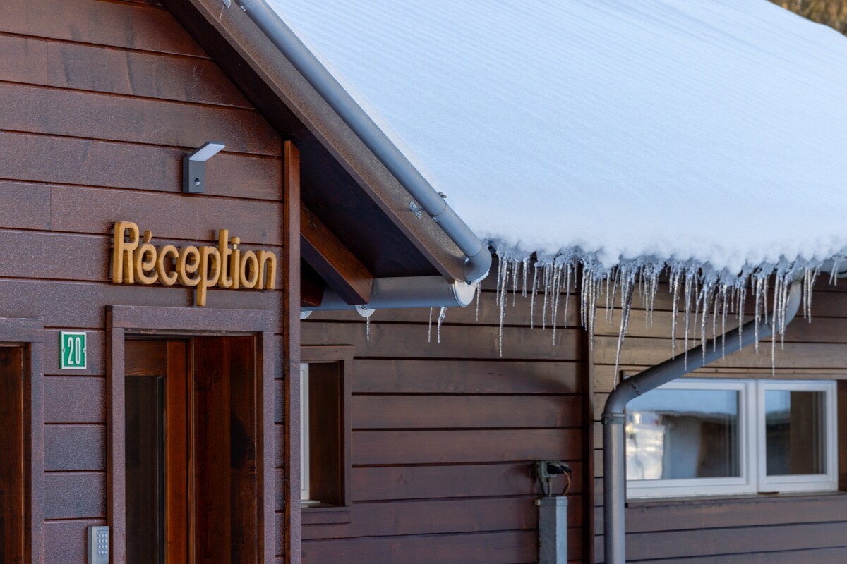 The reception area is depicted with a wooden facade, featuring a sign that reads 'Réception.' Snow accumulates on the roof, with icicles hanging from the edge, suggesting a winter setting. A door is visible, leading into the building.