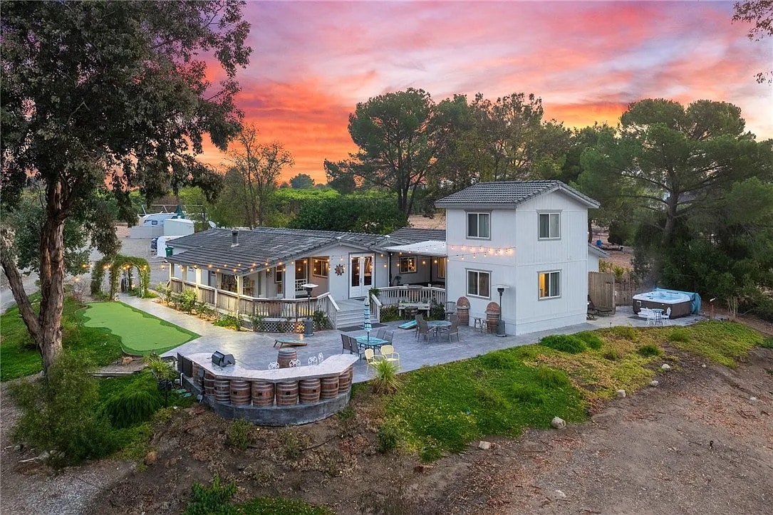 The exterior of the home is captured during sunset, displaying a two-story structure surrounded by trees. A spacious patio area with a hot tub and seating arrangements is visible, complemented by a well-maintained lawn. The vibrant sunset sky adds a warm backdrop to the scene.