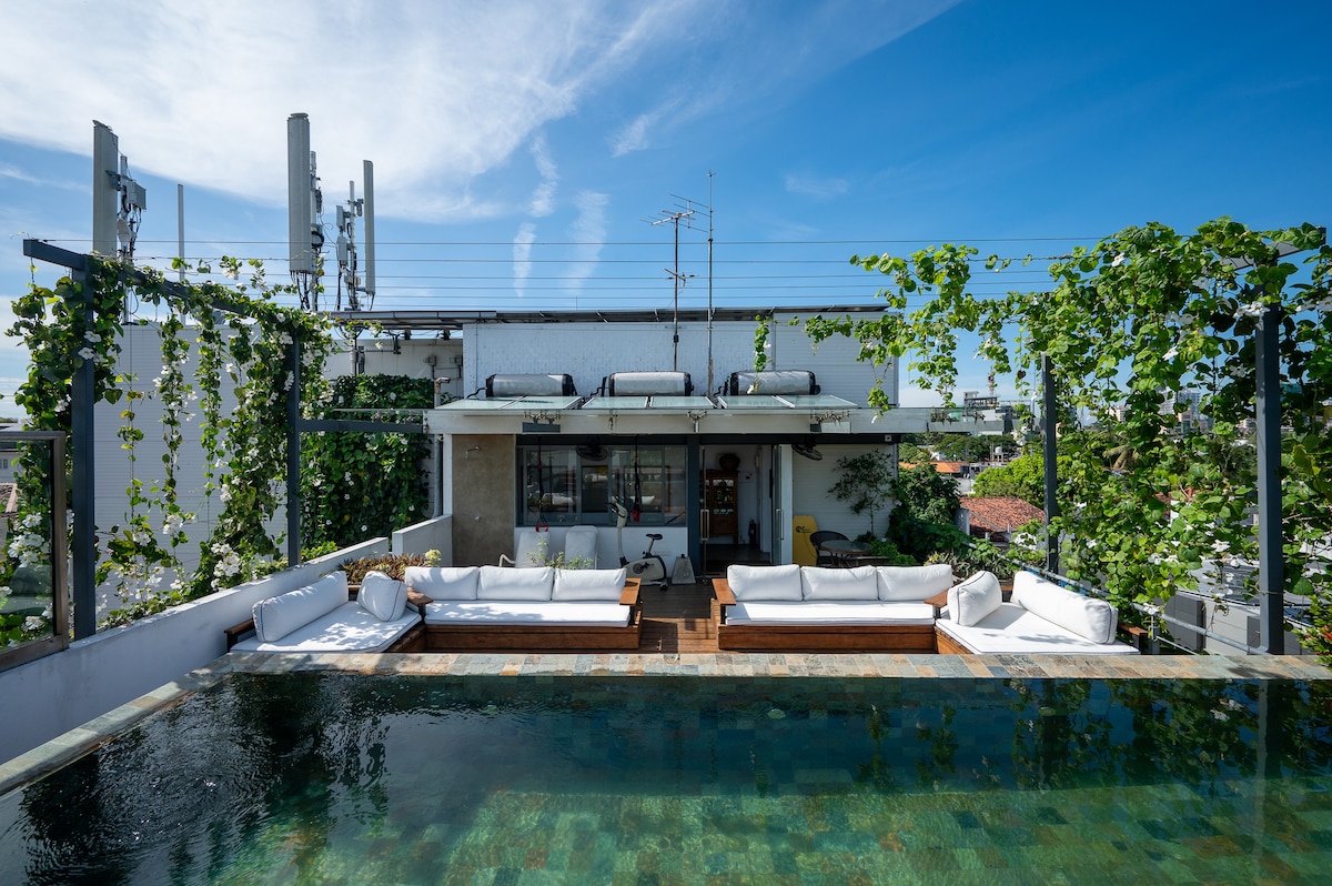 A rooftop terrace features a tranquil swimming pool surrounded by comfortable seating areas. Lush greenery cascades from the edges, enhancing the sense of privacy. Clear blue skies stretch overhead, creating a serene environment for relaxation and leisure.