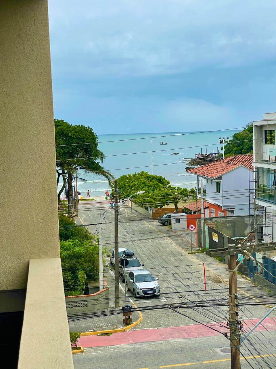 A view from the balcony captures the tranquil beach scene, with sandy shores visible in the distance. The street is lined with trees, and various vehicles can be seen parked along the road. Seagulls fly above the water, enhancing the coastal ambiance.