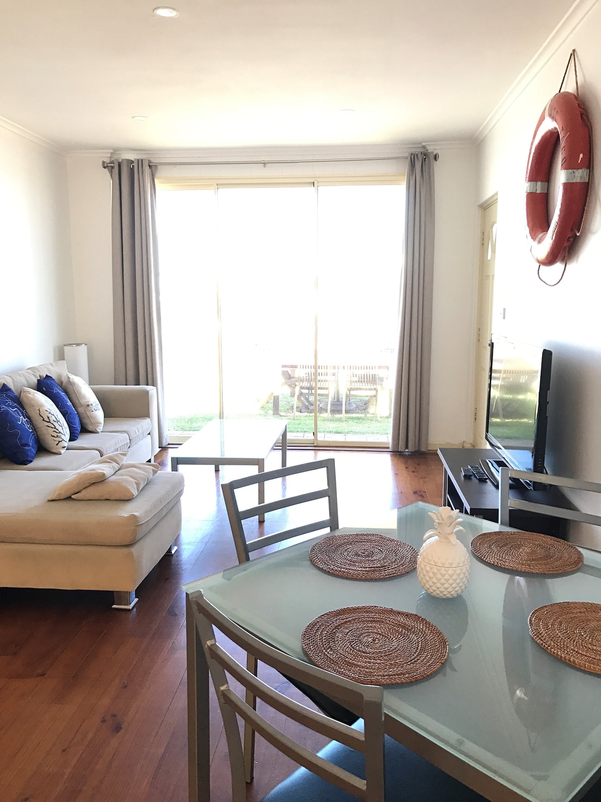 A light-filled living area features a comfortable sectional sofa adorned with decorative pillows. A glass dining table is set with natural placemats. Sliding glass doors offer views of the outdoor space, while a life ring decorates the wall, adding a nautical touch.