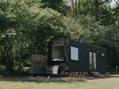 A secluded cabin is nestled among lush greenery, featuring large windows that reflect the surrounding nature. An outdoor copper bathtub is situated next to the entrance, with a wood stack neatly arranged beneath the cabin. A wood burner is visible, enhancing the rustic vibe.