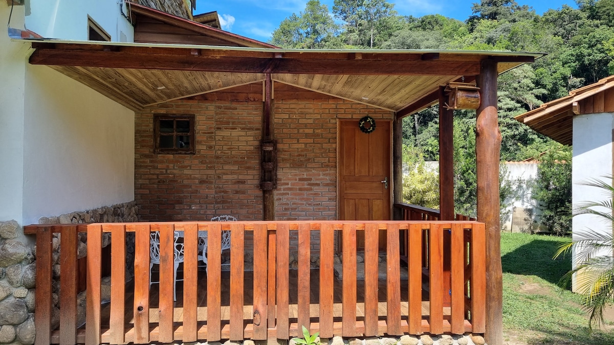 A sheltered porch is visible, featuring a wooden railing and a view of the surrounding greenery. The space includes rustic seating against a brick wall, with natural light filtering through the open area. The entrance door is made of wood, adding to the cozy ambiance.