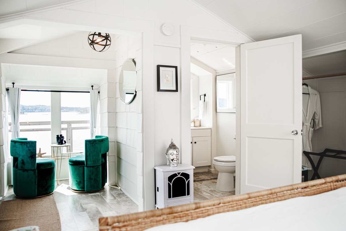 A bright interior is showcased, featuring a cozy sleeping area leading to a seating nook with two green chairs. Natural light floods in through large windows, offering views of the waterfront. A bathroom is visible, equipped with modern fixtures.