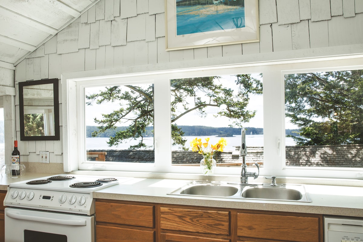 A bright kitchen features large windows that showcase views of the ocean and surrounding trees. The space includes a full stove and double sink, with light-colored cabinetry and a simple decorative flower arrangement positioned by the window.