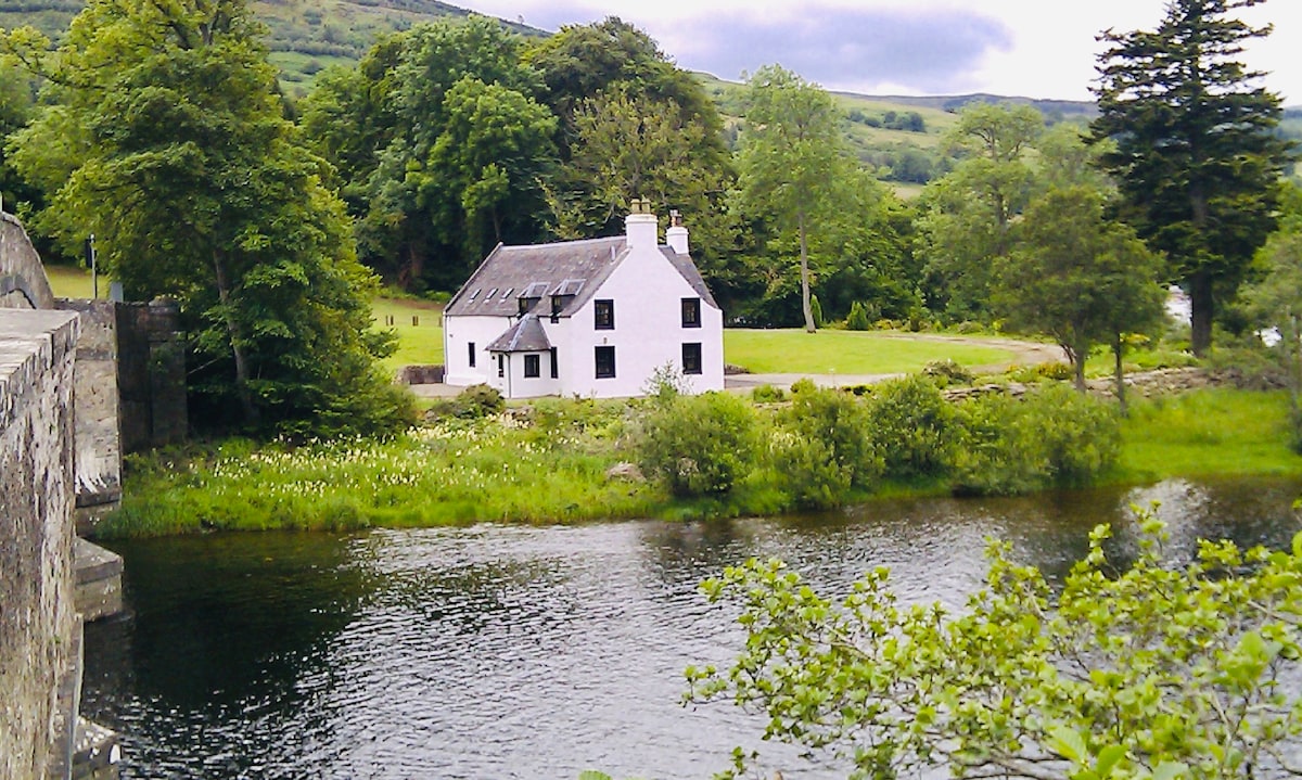 A charming white holiday house sits alongside the serene waters of Loch Tay. Lush greenery surrounds the property, while gentle hills rise in the background. The house features multiple windows that reflect the natural light and provide views of the scenic landscape.