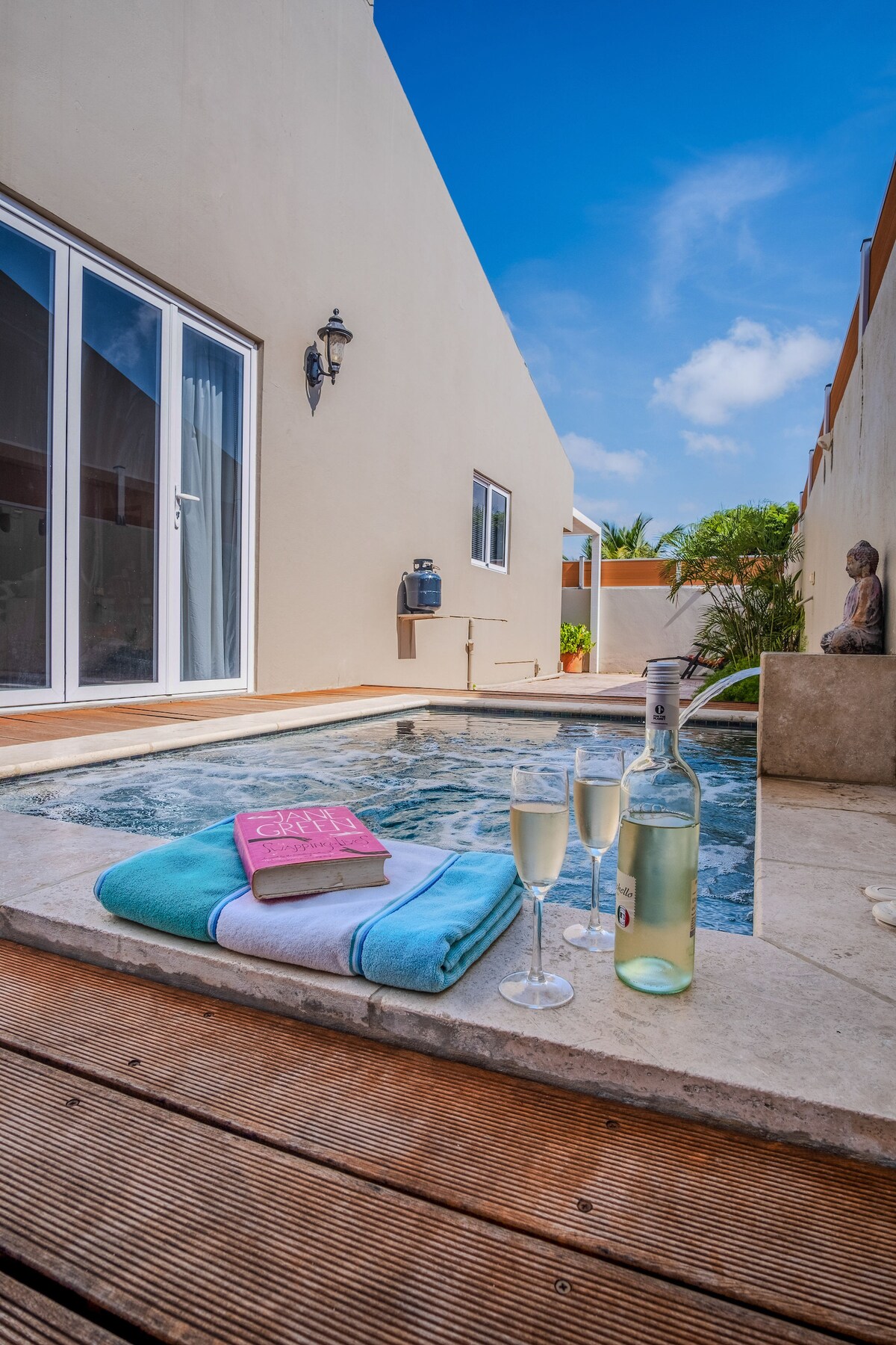 A private plunge pool is set in a sunlit patio area, surrounded by light-colored walls. Two glasses of champagne are placed beside a bottle on a towel, with a book resting nearby. The sky is clear and blue, enhancing the inviting atmosphere.