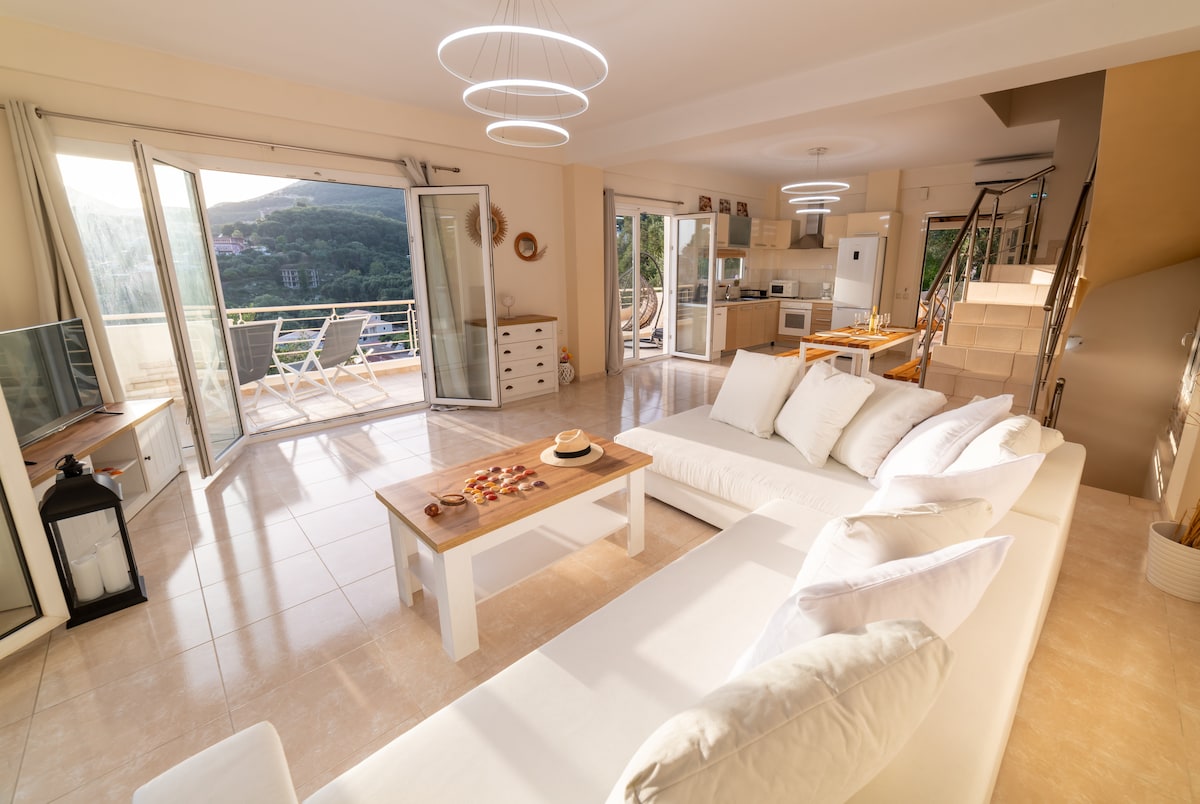An open-plan living area is presented, featuring a large white sofa and a wooden coffee table adorned with snacks. Floor-to-ceiling glass doors lead to an outdoor space, allowing natural light to fill the room and offering scenic views of the surrounding hills.