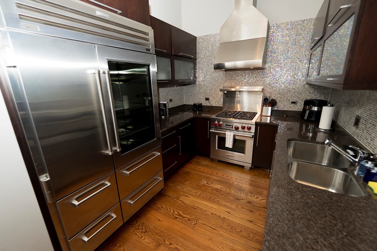 A gourmet kitchen is equipped with professional stainless steel appliances, including a large refrigerator and a sleek gas range with a hood. Mosaic tiles add a modern touch to the wall. The countertop is finished in a dark granite, complemented by warm wooden flooring.