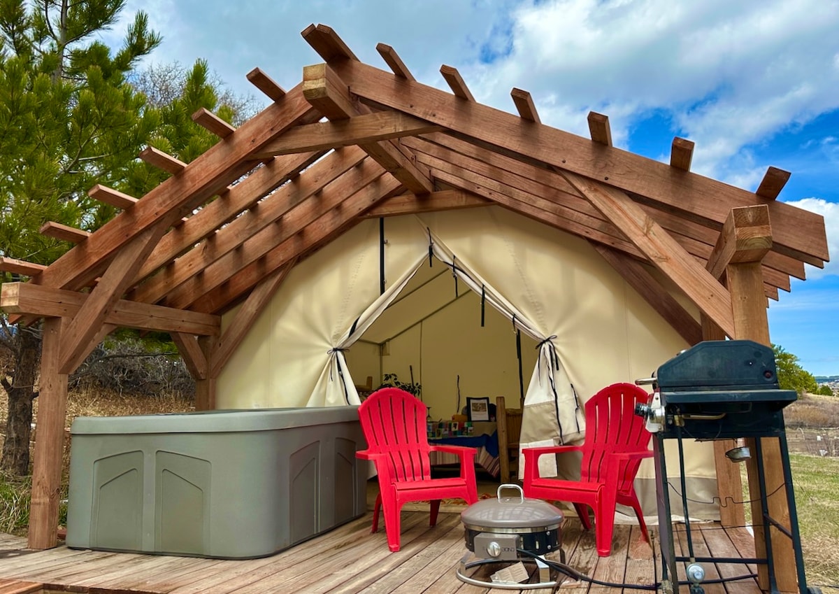 A sturdy wooden structure supports a spacious glamping tent with a friendly entrance. Outside, a hot tub and fire pit are set up on a wooden deck, accompanied by two red lounging chairs and a grill, creating a relaxed outdoor space.