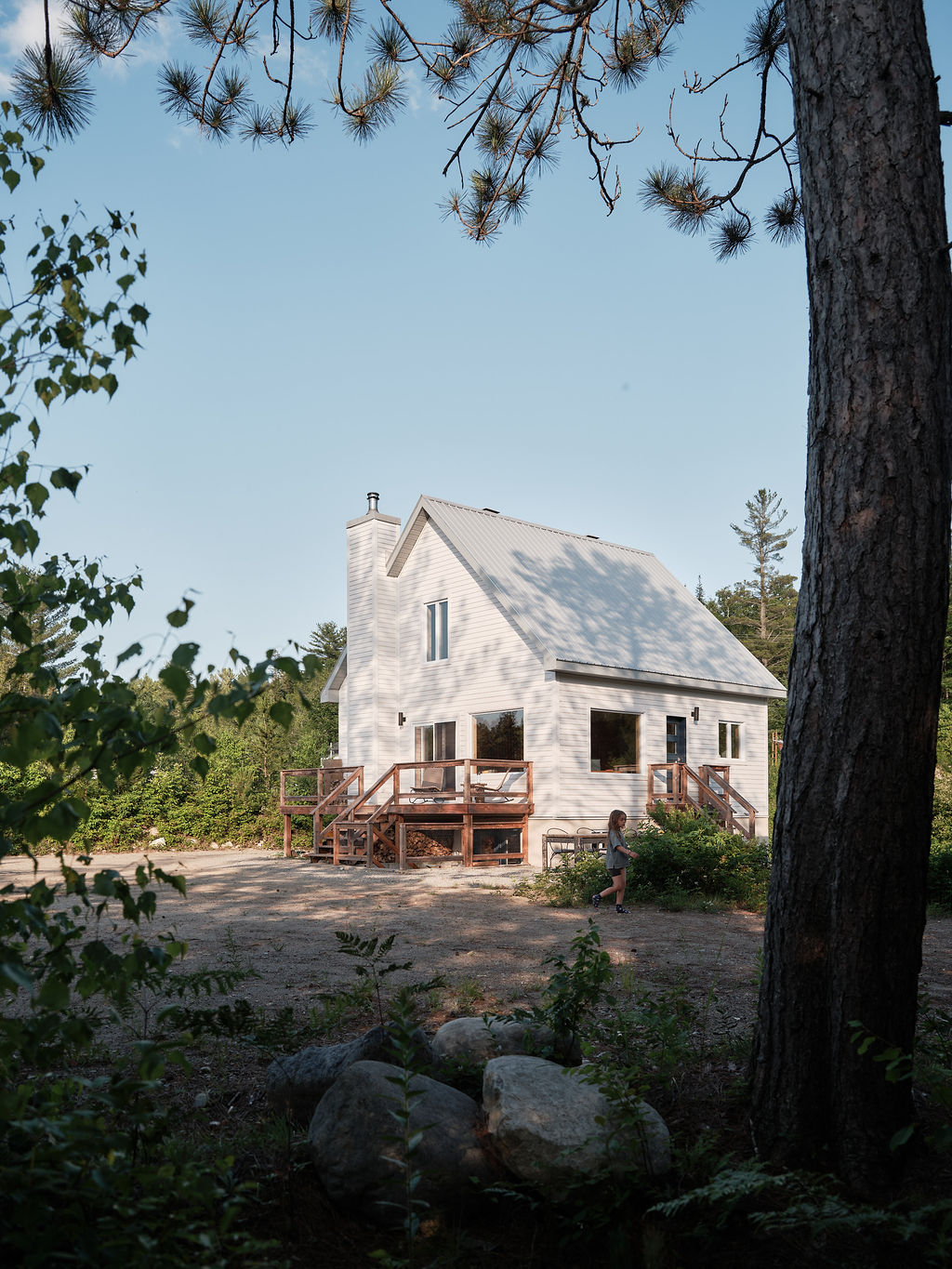 The chalet is nestled among trees, with a wooden deck visible on three sides. Its white exterior reflects the natural surroundings. A gravel area and large boulders are present in the foreground, offering a serene setting amidst the greenery.