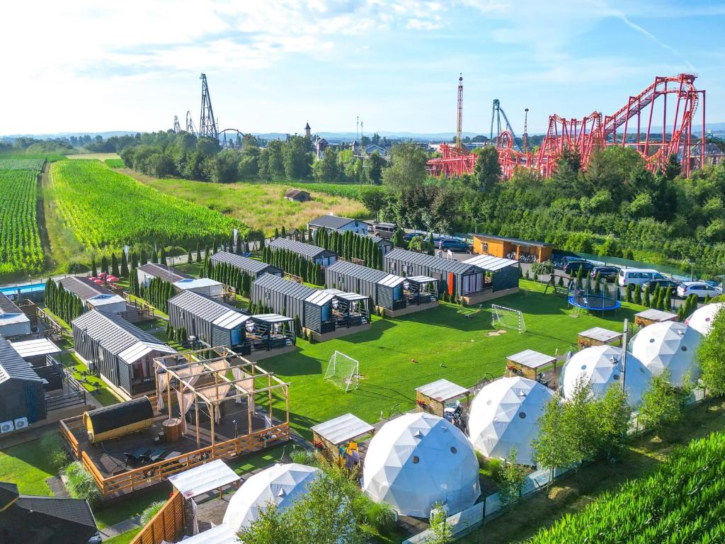 An aerial view of the Holiday Park Zator showcases various accommodation styles, including modern chalets and spherical cabins, set within a lush green landscape. Roller coasters from Energylandia can be seen in the background, surrounded by trees and open spaces.