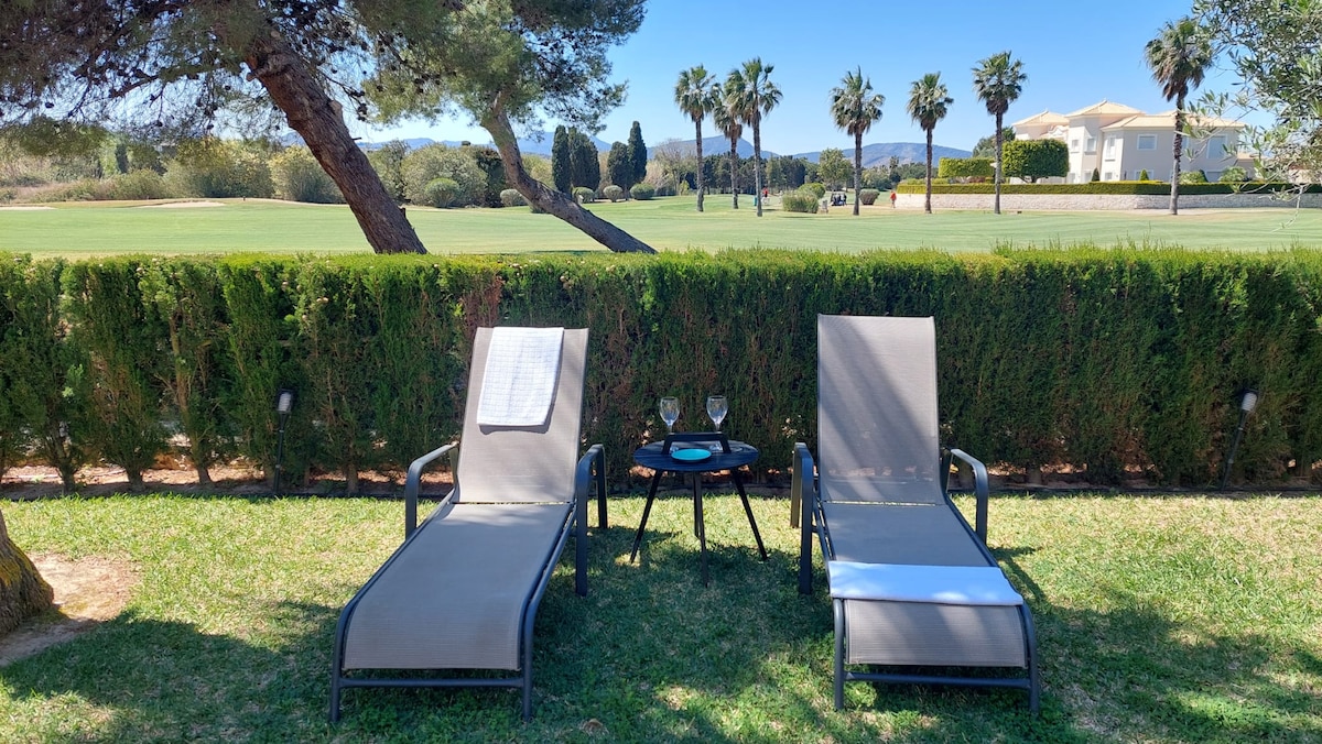 Two lounge chairs are positioned on a grassy area facing a golf course. A small table sits between the chairs, holding two glasses, surrounded by neatly trimmed hedges and palm trees, creating a serene outdoor retreat.