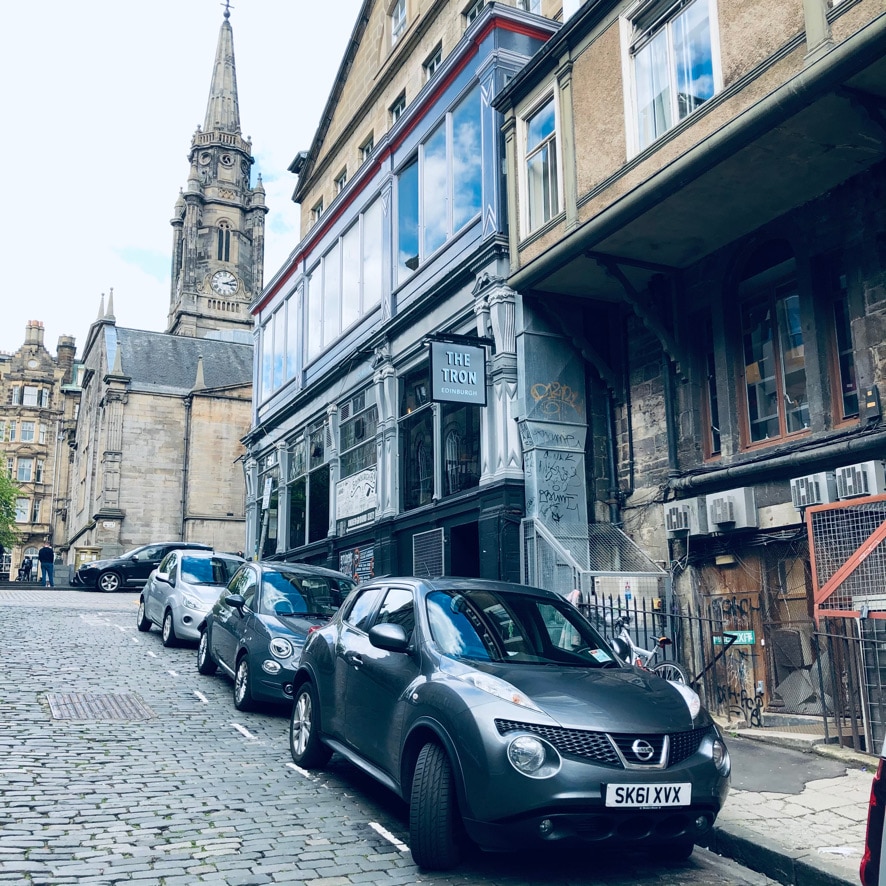 A view of a historic street lined with parked cars, showcasing a blend of modern and older architecture. A distinctive tower can be seen in the background, adding character to the setting. The building features large windows and a sign reading 'The Iron'. Stone paving leads the way.