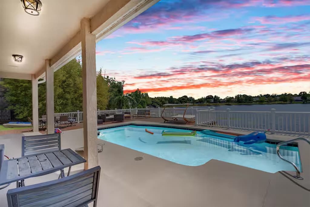 A serene patio space is presented, featuring a private pool with a smooth surface and relaxing lounge items beside it. The view extends over the lake, framed by trees and a colorful sunset sky in the background, enhancing the peaceful environment.