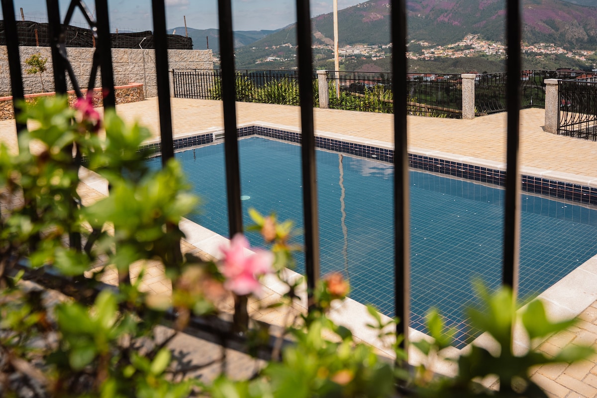 The outdoor swimming pool is framed by a black wrought iron fence, offering a clear view of the serene surroundings. Vibrant flowers add a splash of color in the foreground, while distant mountains are visible in the background, contributing to a peaceful ambiance.