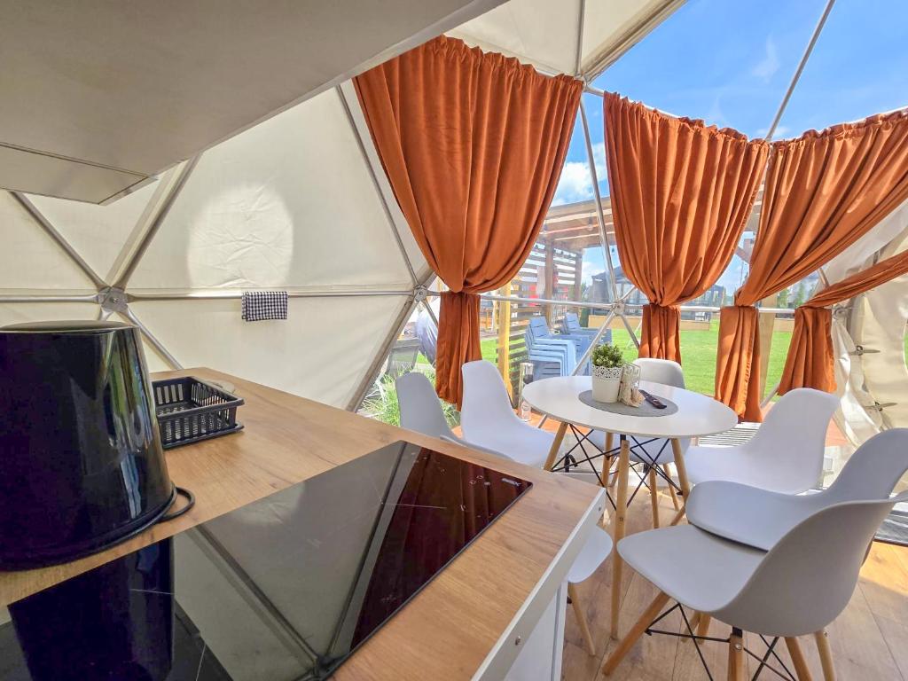 A spacious interior of a spherical cabin features a modern kitchen area with a cooktop and a small black dish rack. A round white dining table is surrounded by four contemporary chairs. Large windows adorned with orange curtains frame the outdoor view of the green garden.