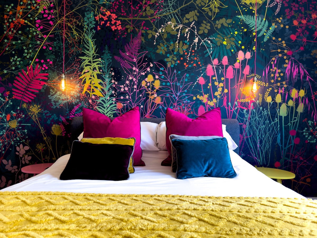 A spacious bed is adorned with an array of colorful pillows in shades of pink, blue, and black. A vibrant floral mural serves as a backdrop, featuring a mix of greens and bright floral patterns. Soft lighting is provided by two hanging light fixtures.