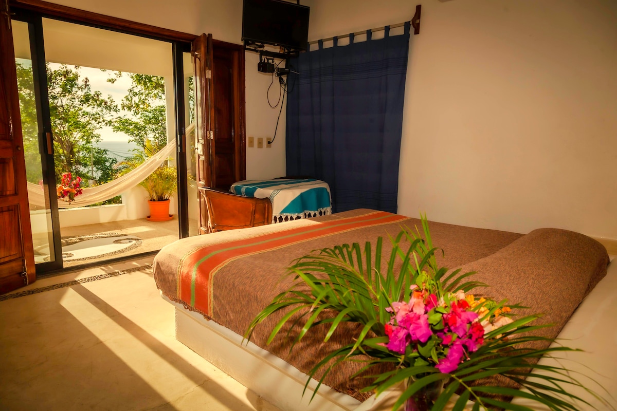 A spacious bedroom features a large bed adorned with natural elements and colorful flowers. A hammock is visible through the open doorway, leading to a balcony area with greenery. Soft light filters through the window, creating a serene and inviting ambiance.