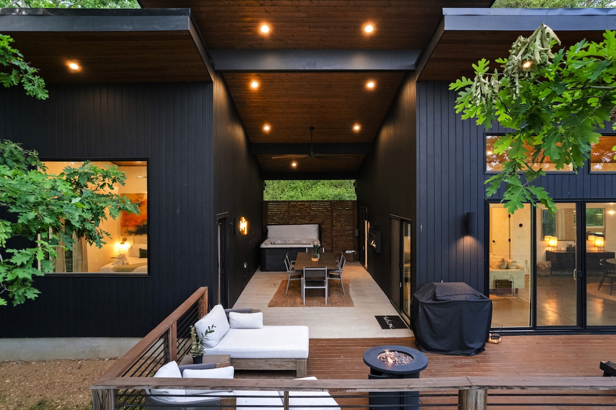 An outdoor living space is highlighted with a spacious deck featuring a seating area, fire pit, and hot tub. The modern structure features large windows and an open design, allowing natural light to illuminate the area. Surrounding trees enhance the private, tranquil setting.