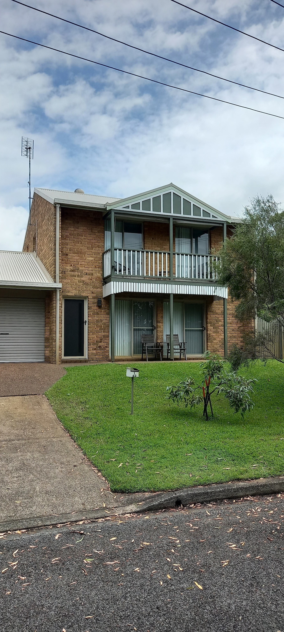 A brick townhouse is pictured, featuring a covered balcony with two chairs and a small table. Lush green grass surrounds the structure, and a driveway leads to a garage. Trees provide a backdrop, while cloudy skies are visible above.